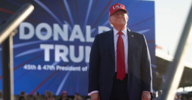 Donald Trump stands on an outdoor stage with his hands by his side while wearing a red baseball cap that has "USA" on it.
