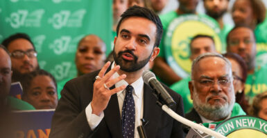 Zohran Mamdani stands and talks into a microphone and gestures with his right hand.
