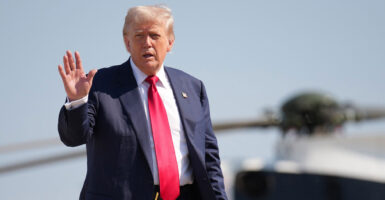 Donald Trump stands and holds his right hand out while a helicopter is in the background.