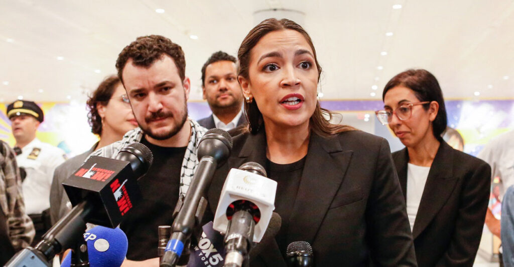 Alexandria Ocasio-Cortez stands in front of a podium and speaks into a microphone as she looks to her left.
