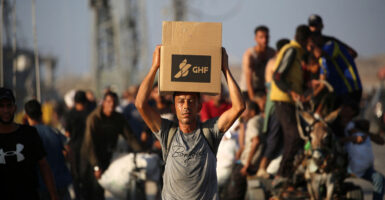 A man in a grey shirt carrying a Gaza Humanitarian Foundation aid parcel on his head walks along a road in the central Gaza Strip.
