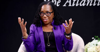 Justice Ketanji Brown Jackson in a purple jacket and black shirt, with her hands lifted, while seated in a white chair.