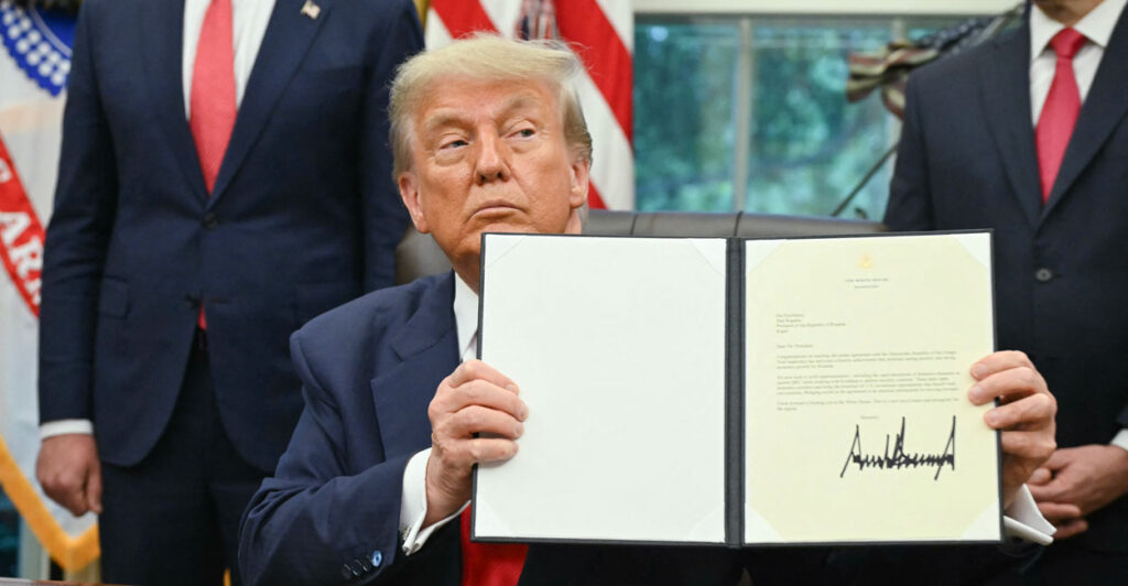 President Donald Trump, sitting at his Oval Office desk, holds up a signed Executive Order.