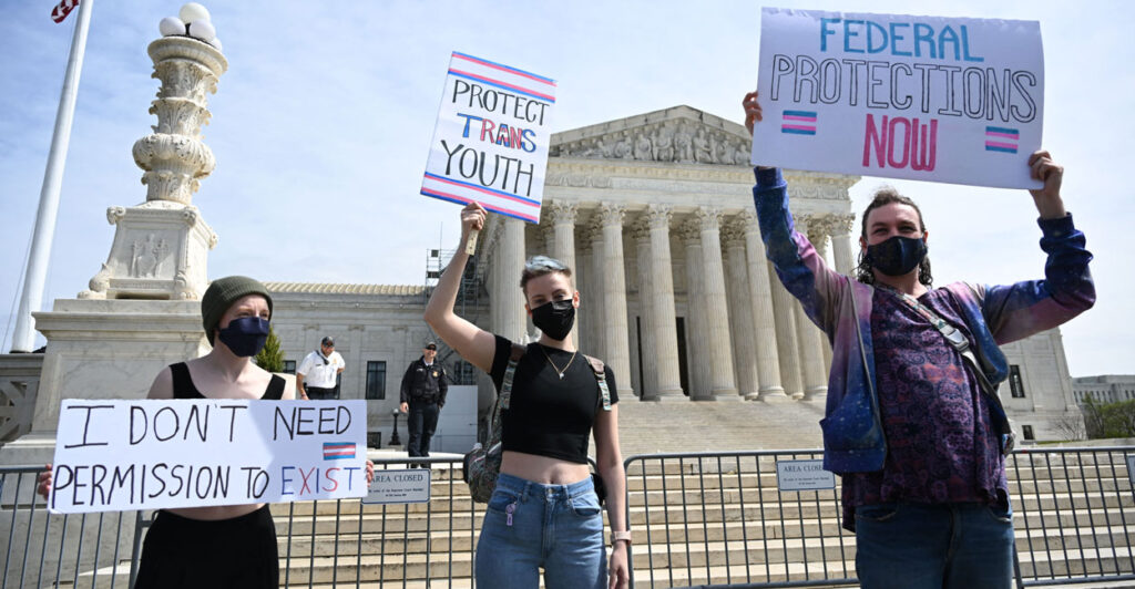 Three trans activists hold signs in front of Supreme Court. One, in jeans and cropped black shirt, holds sign "Protect Trans Youth."