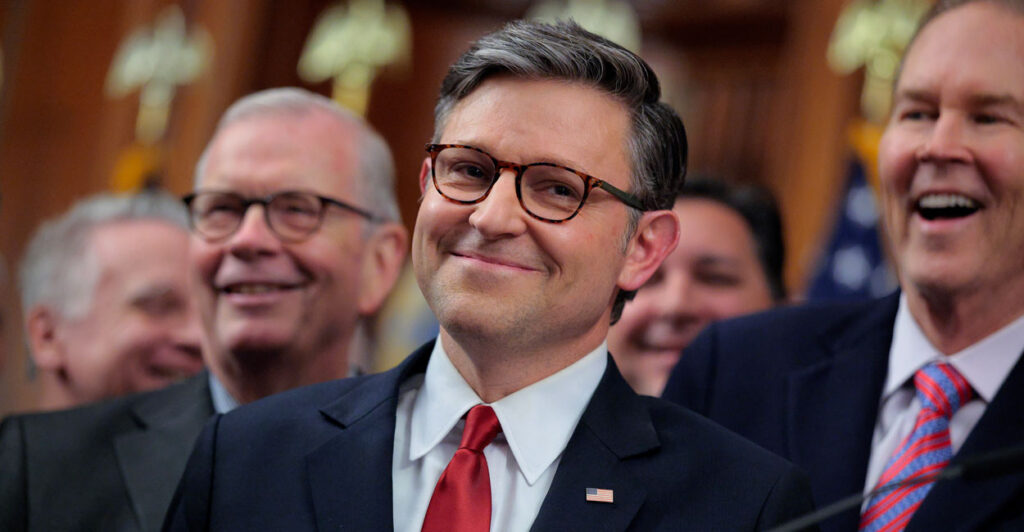 A close up photo of House Speaker Mike Johnson shows him smiling at the camera with other people smiling in the background.