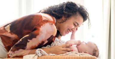 A curly-haired brunette mother leans over top and smiles at her infant, who puts a hand to her face.
