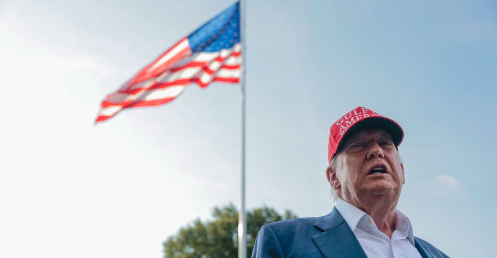 Close-up of President Donald Trump in a MAGA hat, with a flagpole waving an American flag behind him.