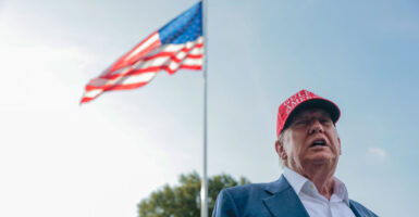 Close-up of President Donald Trump in a MAGA hat, with a flagpole waving an American flag behind him.