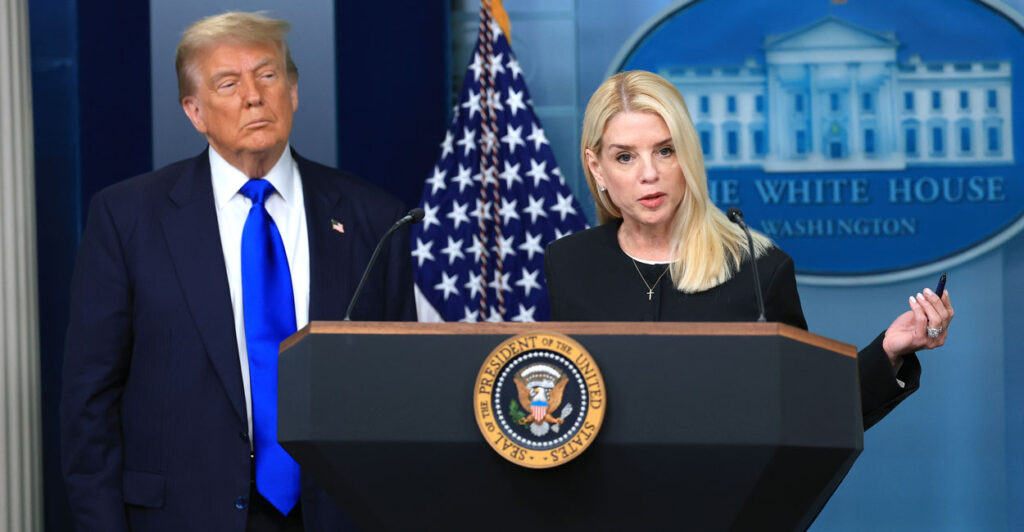 Donald Trump looks on as Pam Bondi, dressed in black, speaks behind a podium in the White House briefing room.