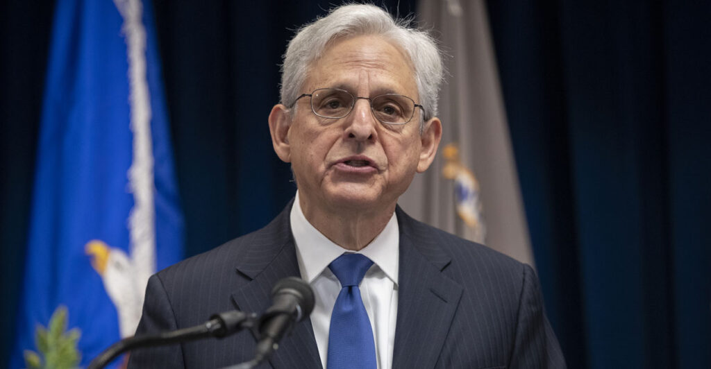 Attorney General Merrick Garland in a dark suit and a blue tie speaks at a podium