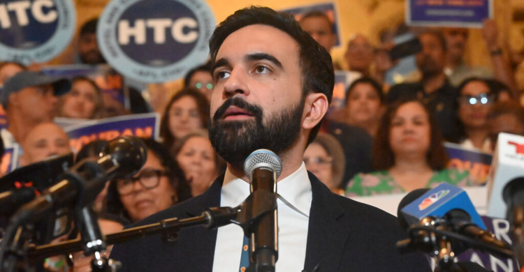 Democratic mayoral candidate Zohran Mamdani lifts his chin as he delivers a speech. Behind him are supporters.