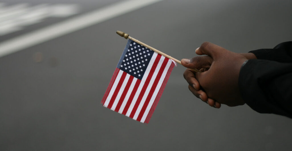 Black hands hold a small American flag next to a street.