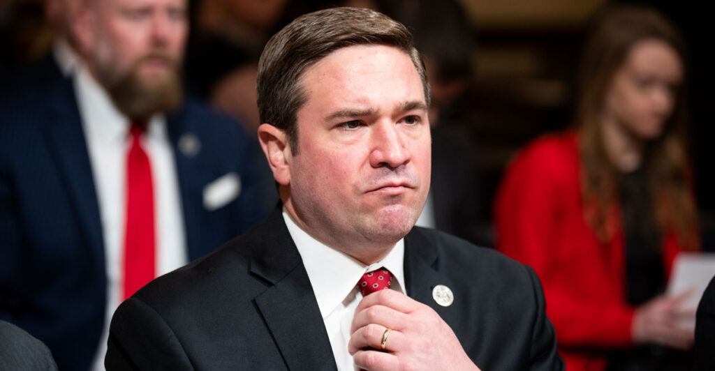 Missouri Attorney General Andrew Bailey sits during a congressional hearing, wearing a black suit jacket and adjusting his red tie.