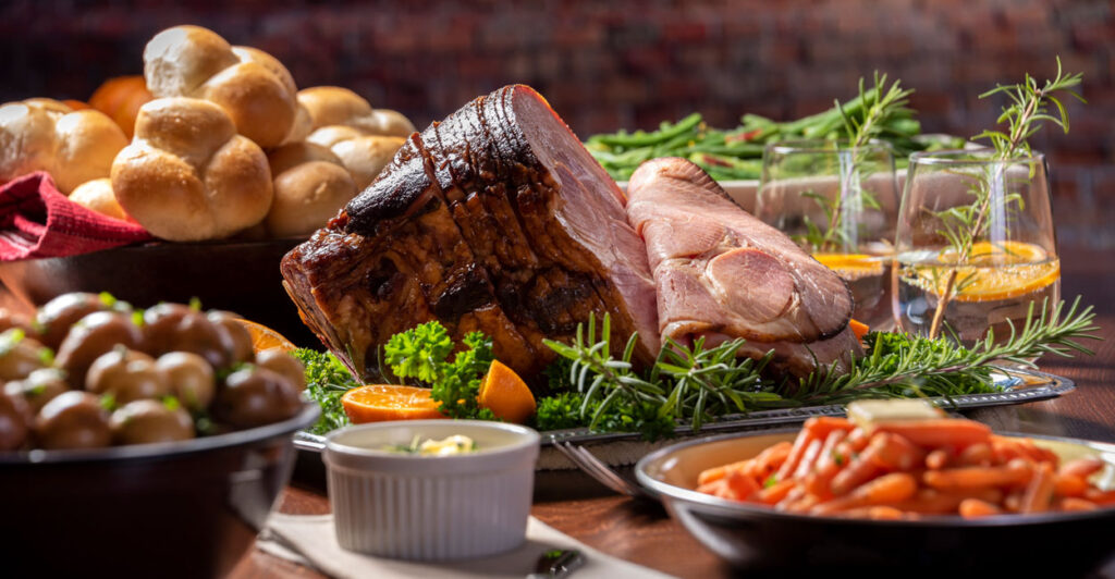 A mouth watering ham dinner, with bread, carrots, potatoes and greens joining a glazed ham.