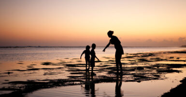 A mother and her two kids in silhouette at sunset their feet in the ocean.