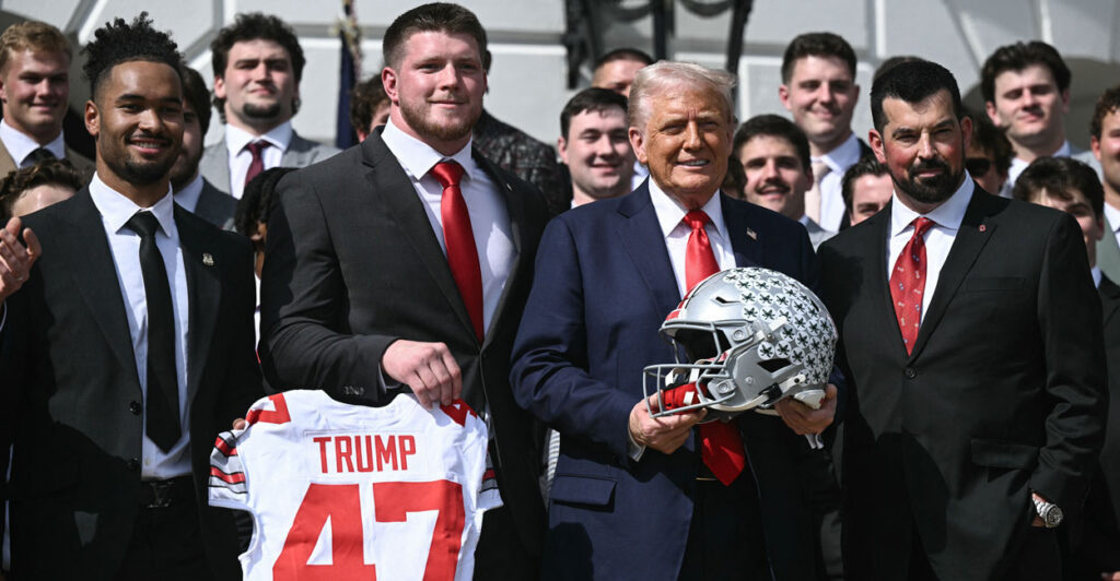 President Donald Trump holds an Ohio State football helmet while surrounded by the NCAA championship football team in front of the White House.