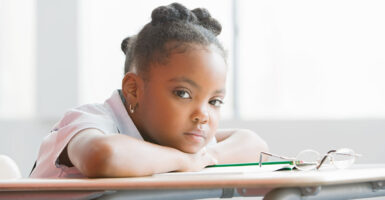 An intense black school girl rests her head on her hands and desk, staring at the camera.