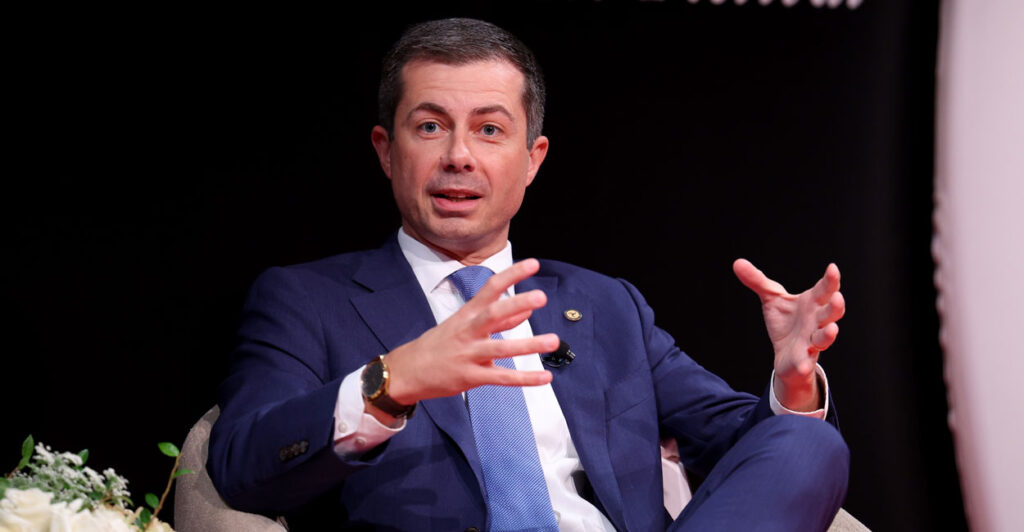 Former transportation Secretary Pete Buttigieg In a blue suit and light blue tie speaks to an audience and gestures with his hands