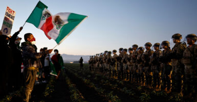 Protesters waving Mexican flags confront a line of National Guard soldiers near a marijuana farm as the sun fades.