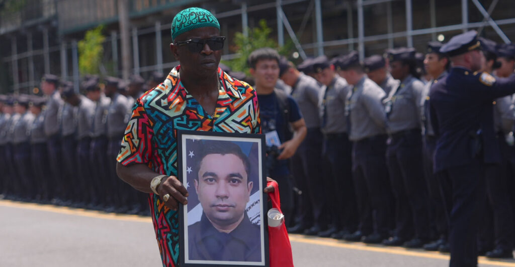 A man in colorful shirt holds a picture of slain NYPD officer Didarul Islam during his funeral procession. Along the street a line of officers bow their heads.