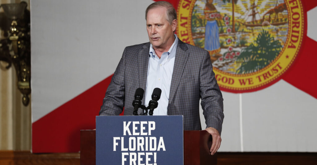 Florida Agriculture Commissioner Wilt Simpson in a suit jacket stands at a podium with the sign that says keep Florida free