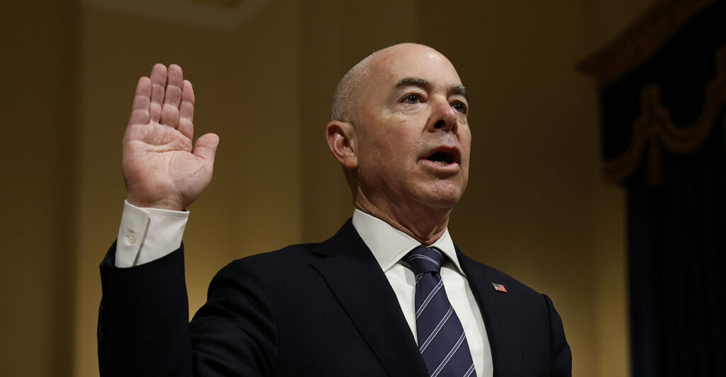 Alejandro Mayorkas in a black suit raising his hand.