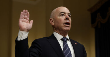 Alejandro Mayorkas in a black suit raising his hand.