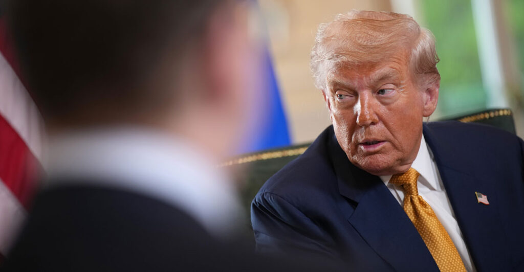 President Donald Trump is visible beyond a man's shoulder. Trump wears a golden yellow tie and is turned slightly as he speaks to someone.