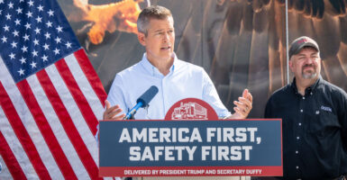 Transportation Secretary Sean Duffy In a collared shirt speaks Outdoors at a podium that has a sign that says America first, safety first