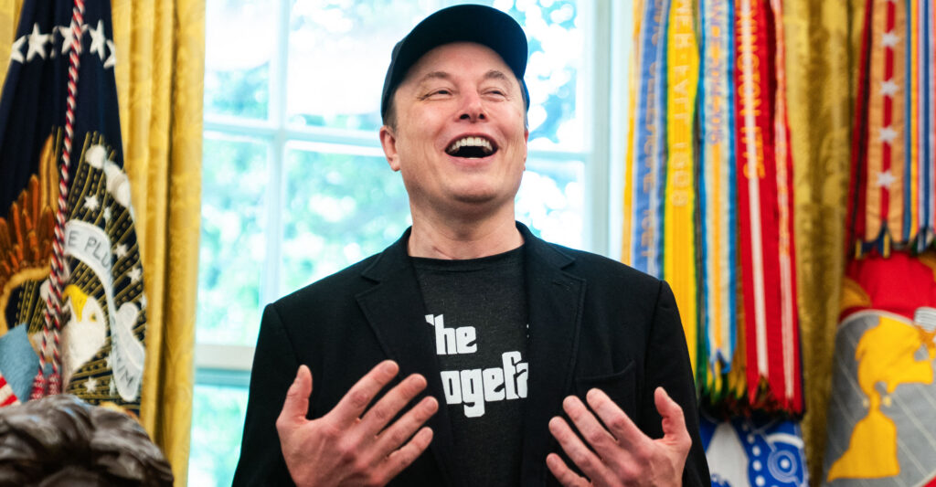 Elon Musk, in a dark jacket and black T-shirt, stands in the Oval Office with his mouth wide, hands at his chest.