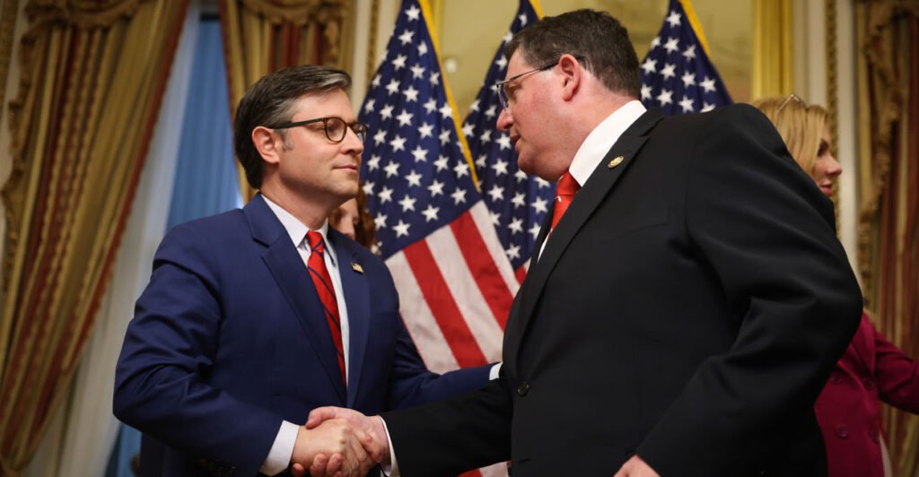 House Speaker Mike Johnson and Rep. Randy Fine shake hands in front of American flags.