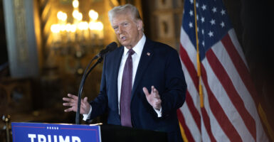 President Donald Trump in a dark suit speaks at a podium at his Mar-a-Lago Club. A US flag is in the background