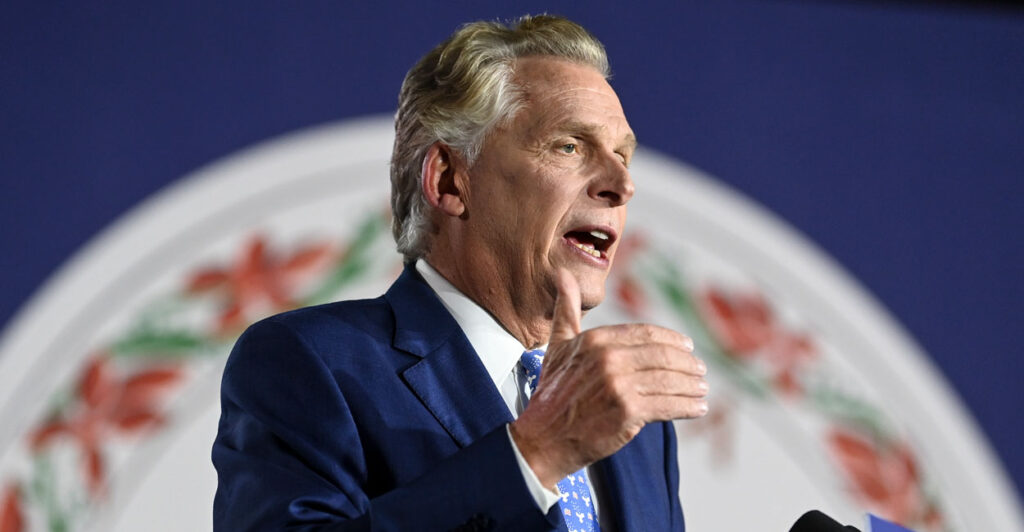 Former Virginia Governor Terry McAuliffe in a dark suit gesturing as he speaks at a podium with the seal of Virginia in the background