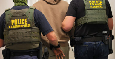 Two border patrol agents detain a man wearing a tan hoodie.