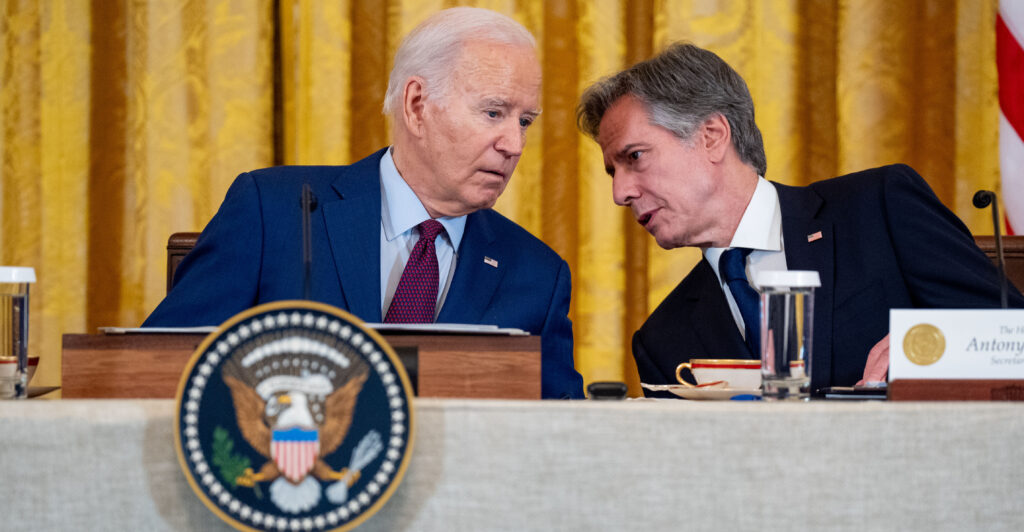 Then-President Joe Biden speaks with then-Secretary of State Antony Blinken as both are seated at a table behind the presidential seal