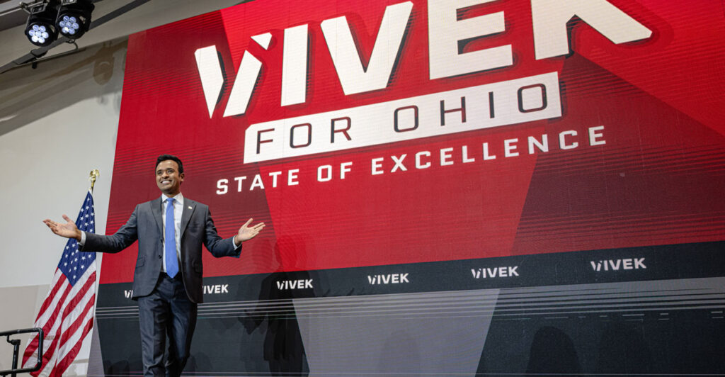 Vivek Ramaswamy gestures to the crowd as he stands in front of a billboard from his campaign.