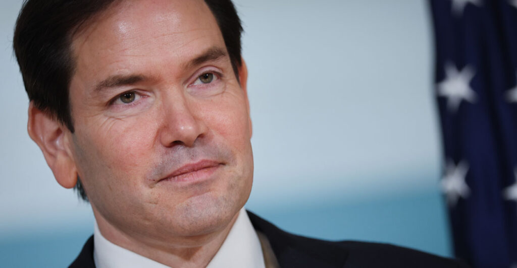 A close up photo shows Secretary of State Marco Rubio in a suit with an American flag over his shoulder.