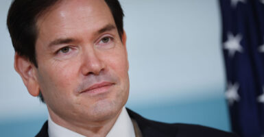 A close up photo shows Secretary of State Marco Rubio in a suit with an American flag over his shoulder.