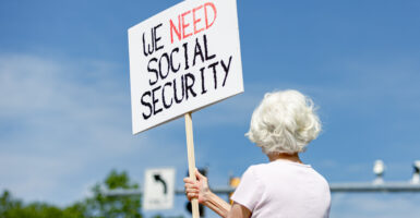 A woman holds a handwritten sign up in public that says "we need Social Security"