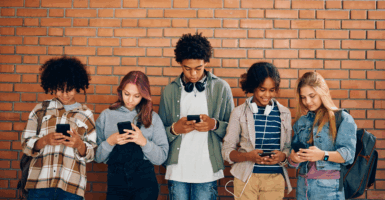 A group of kids stand in front of a brick wall looking at their phones