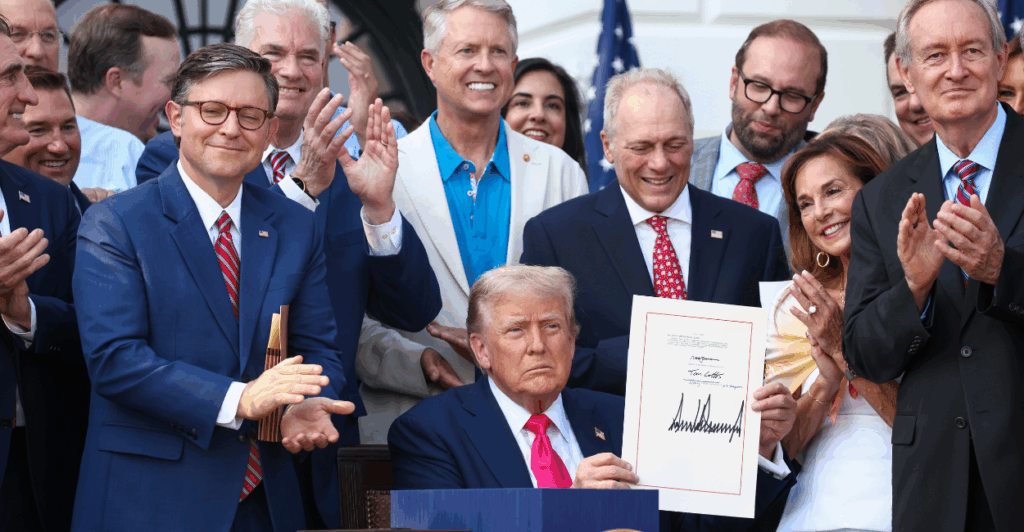 President Donald Trump, wearing a navy suit and red tie, holds up a signed bill while GOP lawmakers applaud