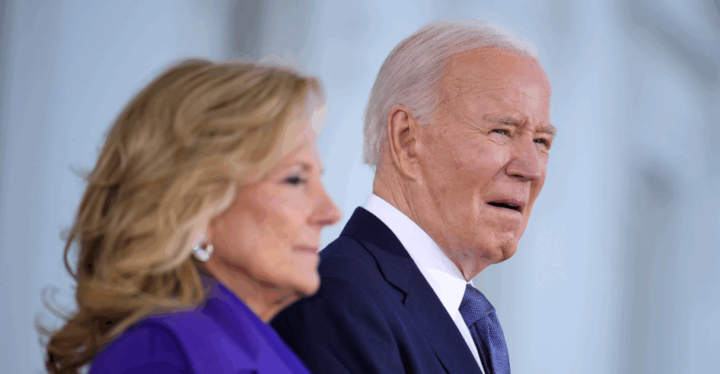 Jill Biden in a purple suit stands in the foreground; in the background, President Joe Biden, wearing a navy suit and tie, squints ahead.
