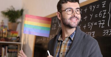 A male teacher holding gay pride flag in front of a chalkboard.