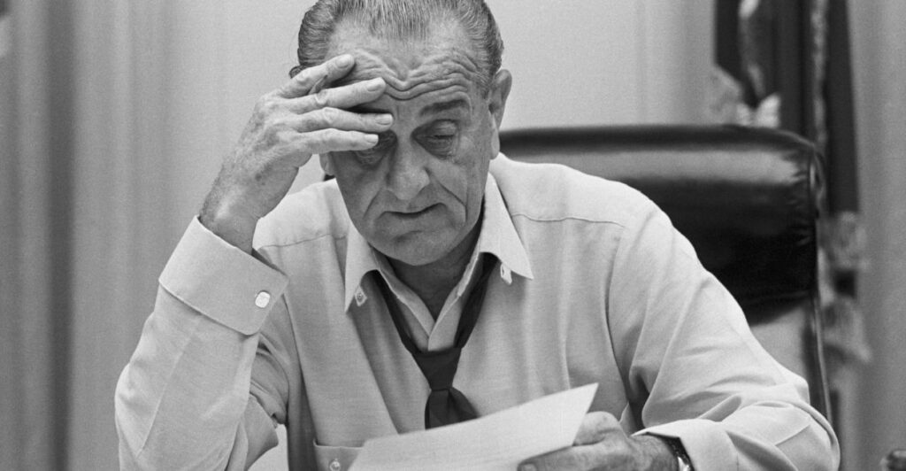 President Lyndon B. Johnson sits looking at a paper, which his hand on his forehead.