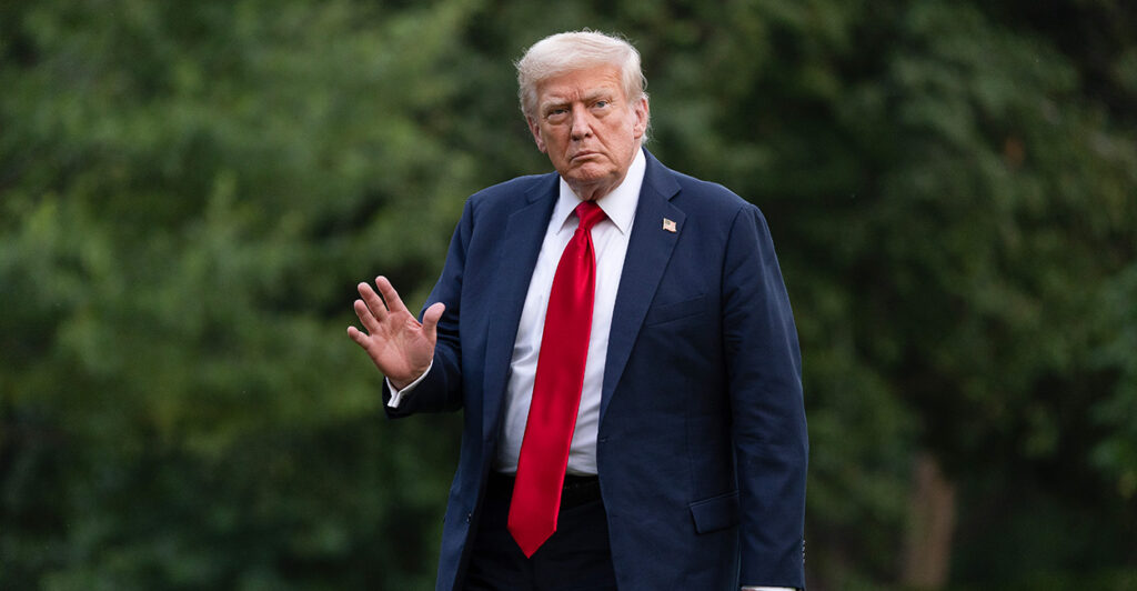 President Donald Trump in a blue suit and red tie waving as he walks outside the White House.