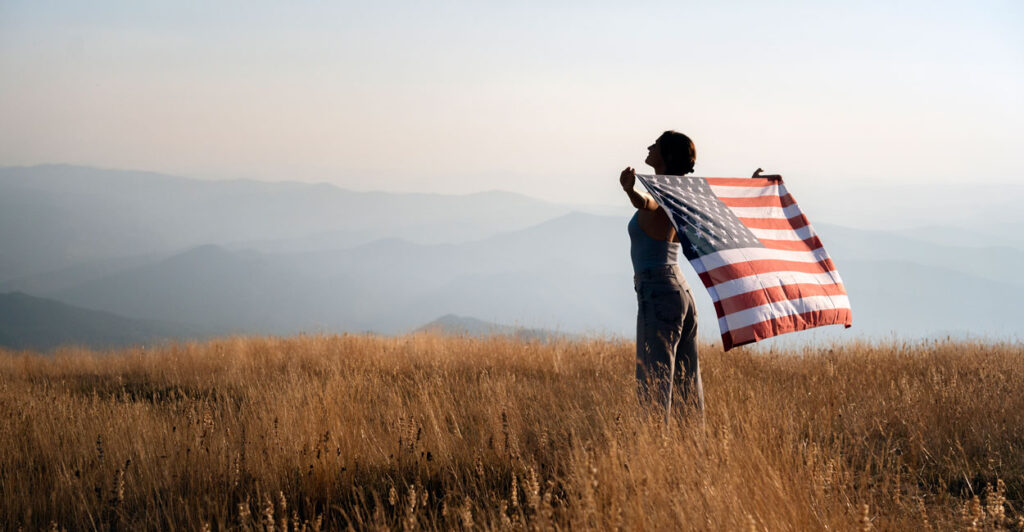 a woman in a field of tall grass overlooking a mountain range below holding an American flag in her arms