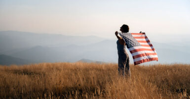 a woman in a field of tall grass overlooking a mountain range below holding an American flag in her arms