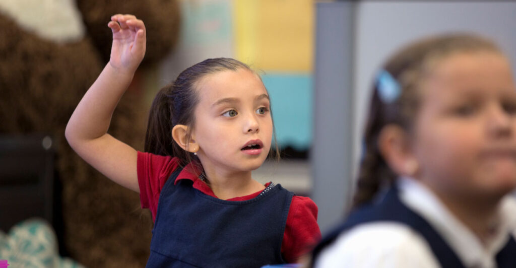 Cute little girl in red blouse over a blue dress raises her hand. Classmate in school uniform, slightly out of focus, in the foreground.