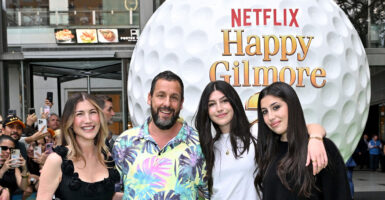 Adam Sandler stands in front of a promotional poster with his wife and two daughters on the red carpet.