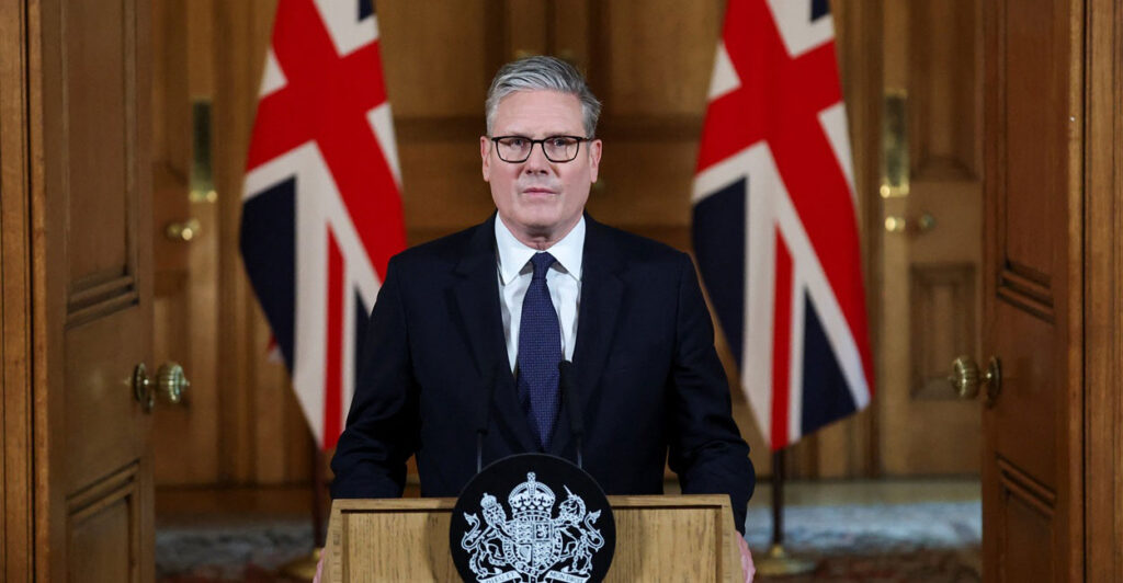 Keir Starmer stands behind a podium and looks straight ahead with two British flags behind him.